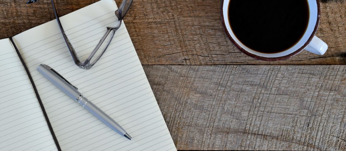A journal, pen, glasses, and mug of coffee